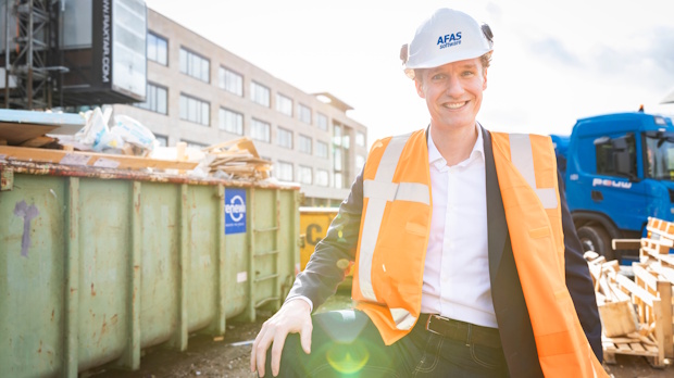 Man wearing a white hard hat and orange safety vest stands at a construction site with green dumpsters and construction materials in the background, smiling at the camera.