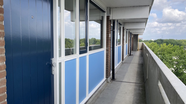 Long exterior corridor of an apartment building with blue doors and large windows, brick columns, and a railing overlooking green trees.