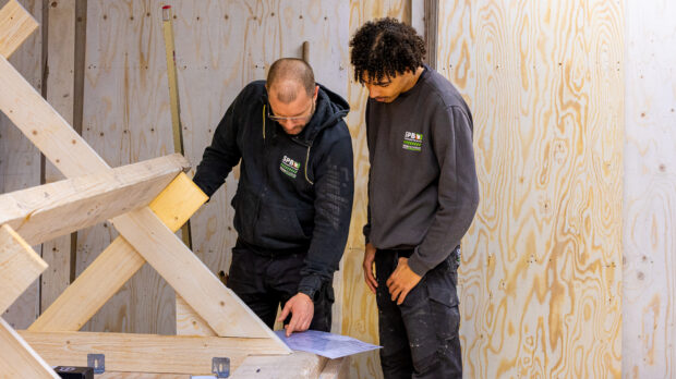 Two construction workers in dark clothing study blueprints on a wooden worktable inside an unfinished building with plywood walls and scattered framing lumber.