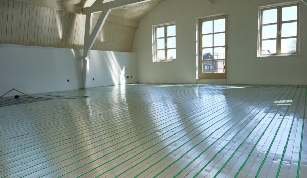 Sunlit unfinished room with exposed beams and large windows; radiant floor heating panels with green tubing installed across the floor.