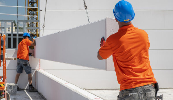 Two construction workers in orange shirts and blue hard hats guide a large white panel into place with a crane.