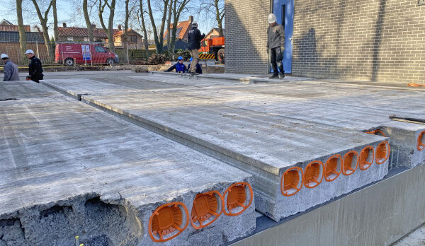 Construction site with large concrete slabs and orange edge inserts, workers nearby beside a brick building with a blue door in the background.