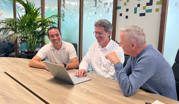 Three colleagues at a wooden conference table in a bright office, smiling at a laptop screen as they discuss work.